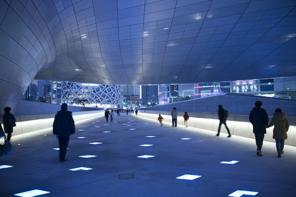 People walking along illuminated walkway underneath curved ceiling of contemporary building.